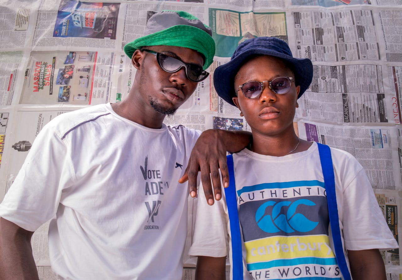 Home Fashionable photo of two men in sunglasses and hats, posing confidently against a newspaper backdrop in Nigeria.